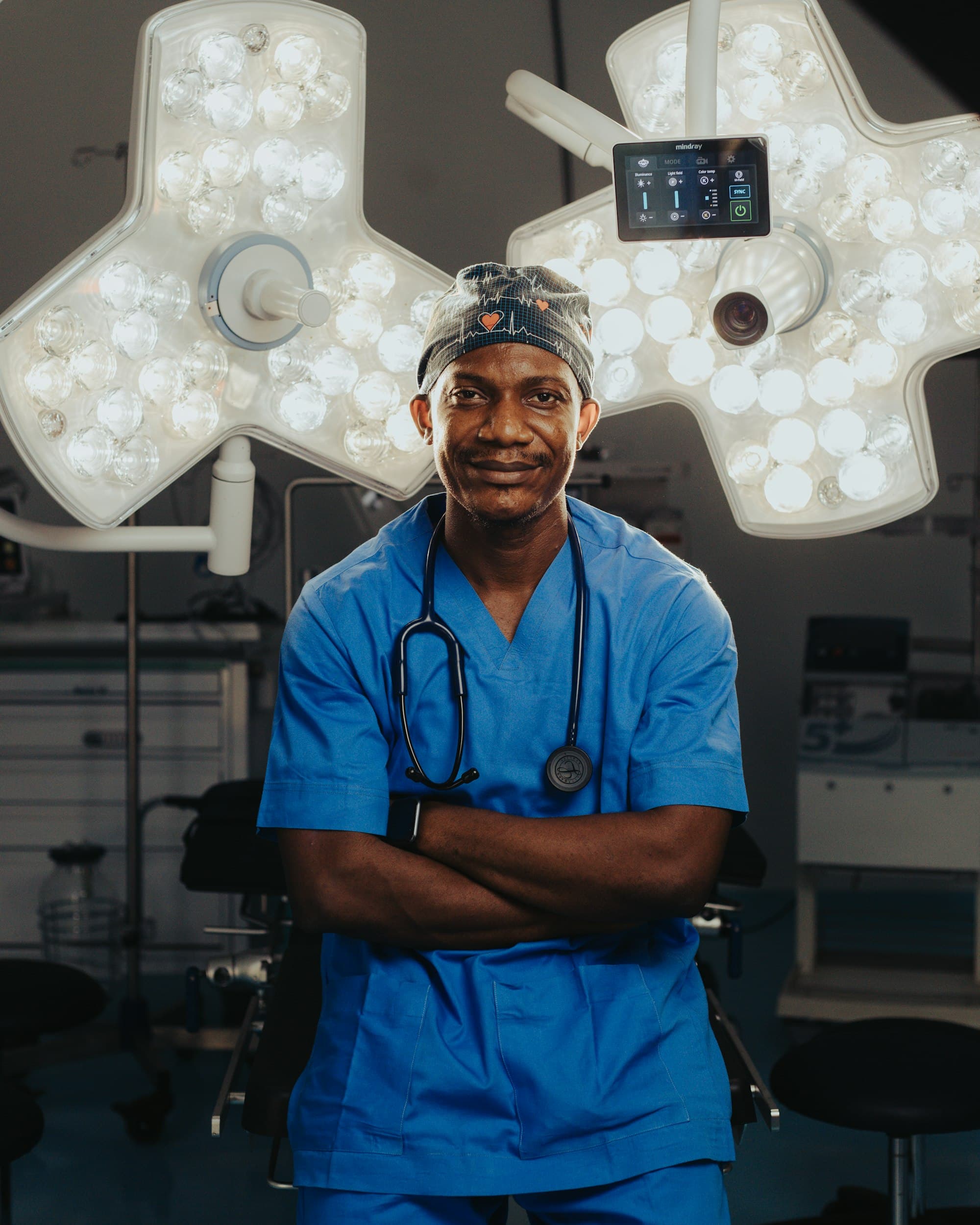 A surgeon in blue scrubs standing under theatre lights, arms folded.