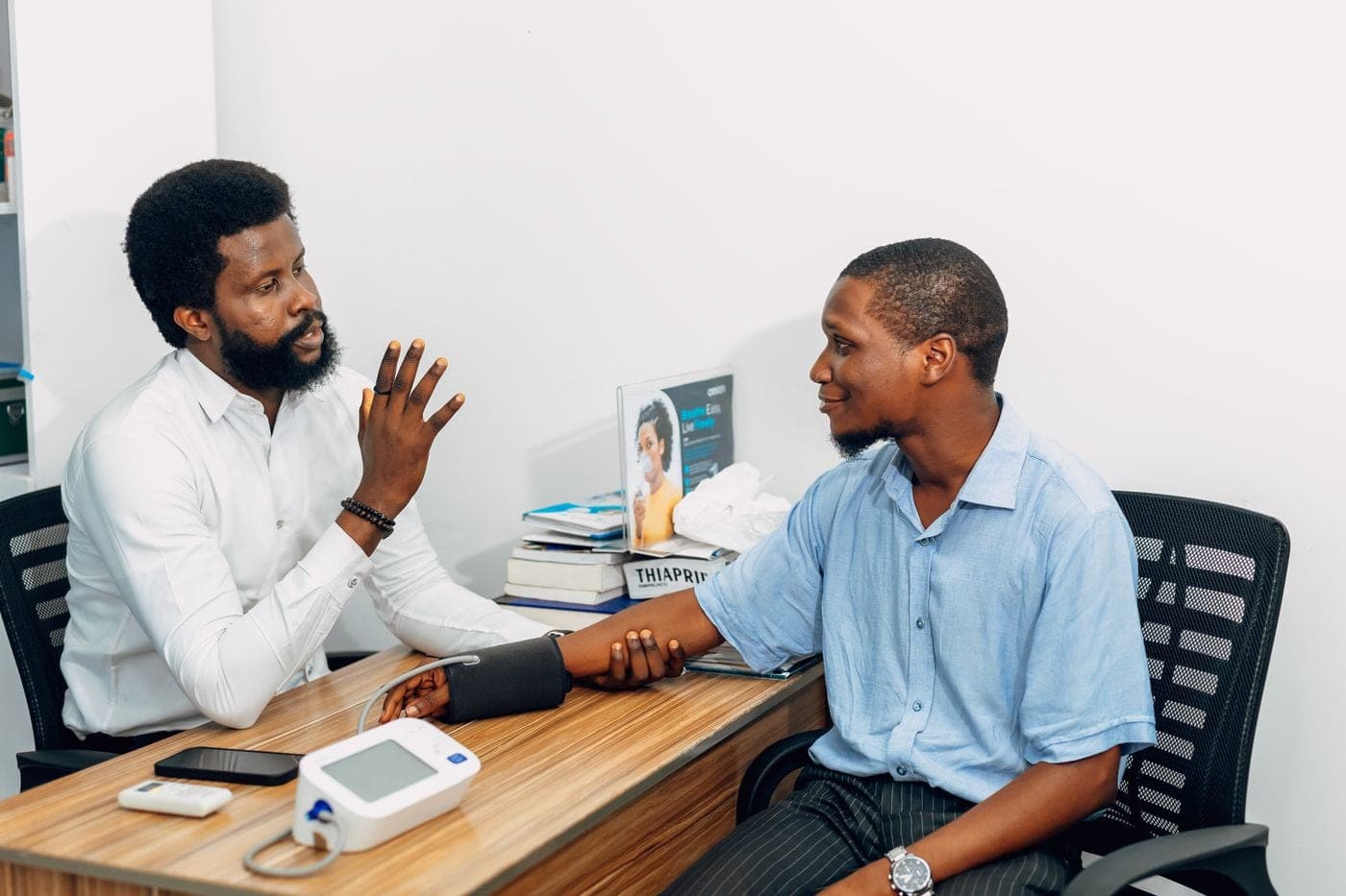 A doctor talking with a patient across a clinic desk, a blood pressure monitor between them.