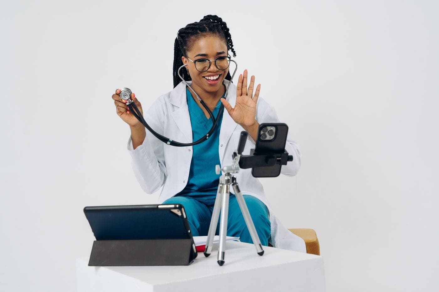 A smiling doctor waving to a patient on a video call, holding up her stethoscope.